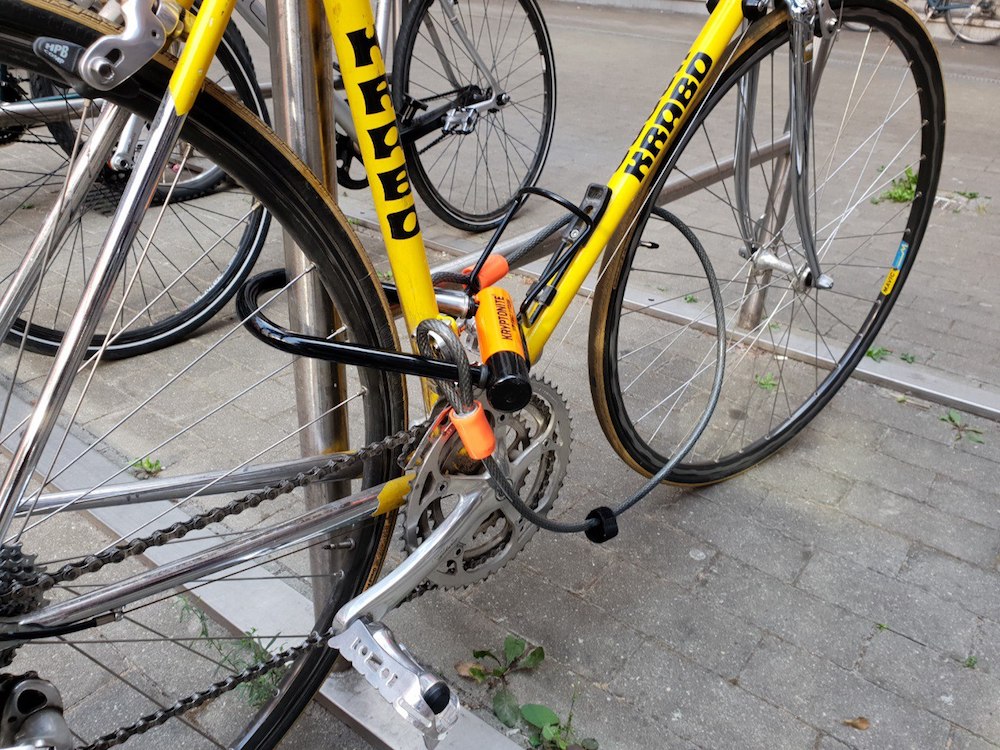 Yellow bike in a parking space locked with a U-lock and a flexible steel cable.